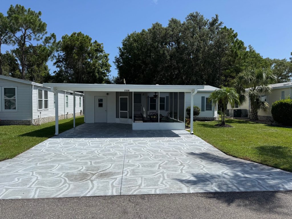 A white manufactured home with a covered carport, screened porch, and patterned concrete driveway, situated between similar homes and surrounded by green grass and trees.