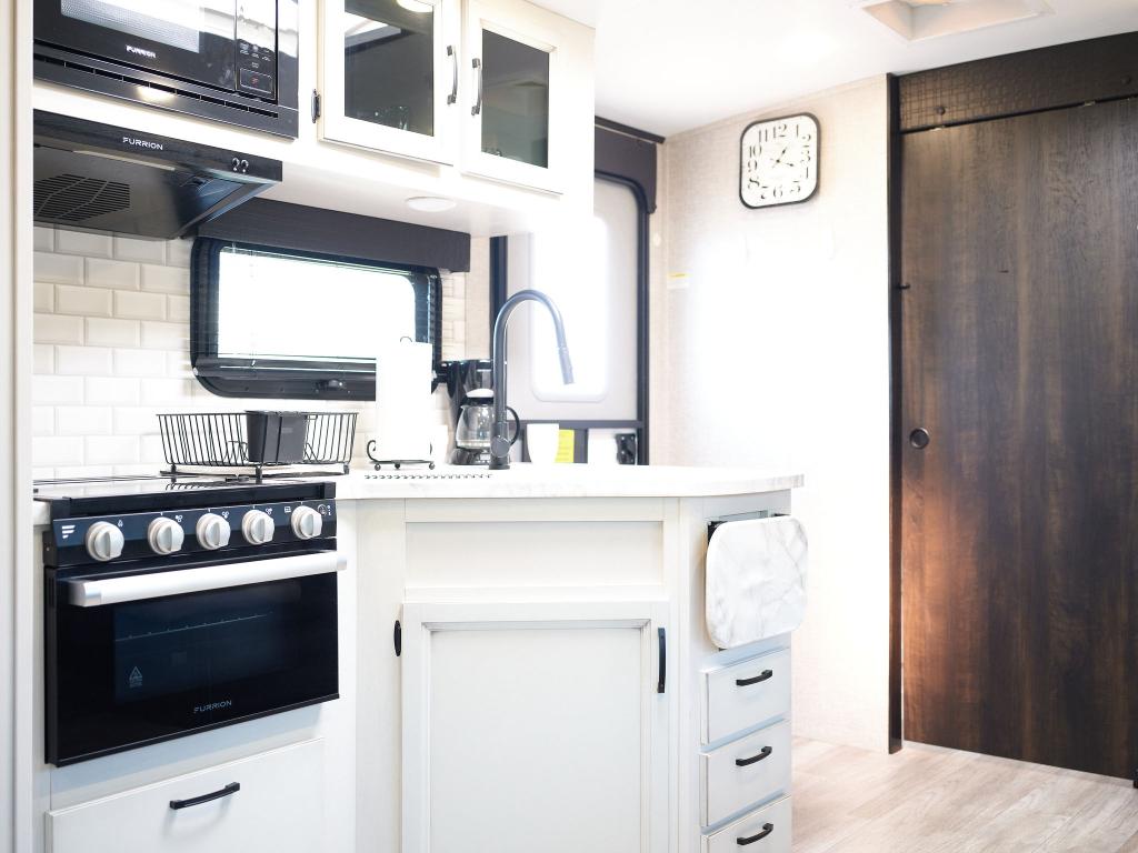 Interior of a modern RV kitchen featuring a stove, white cabinets, black microwave, faucet, and a wall clock. A sliding wooden door is visible on the right.