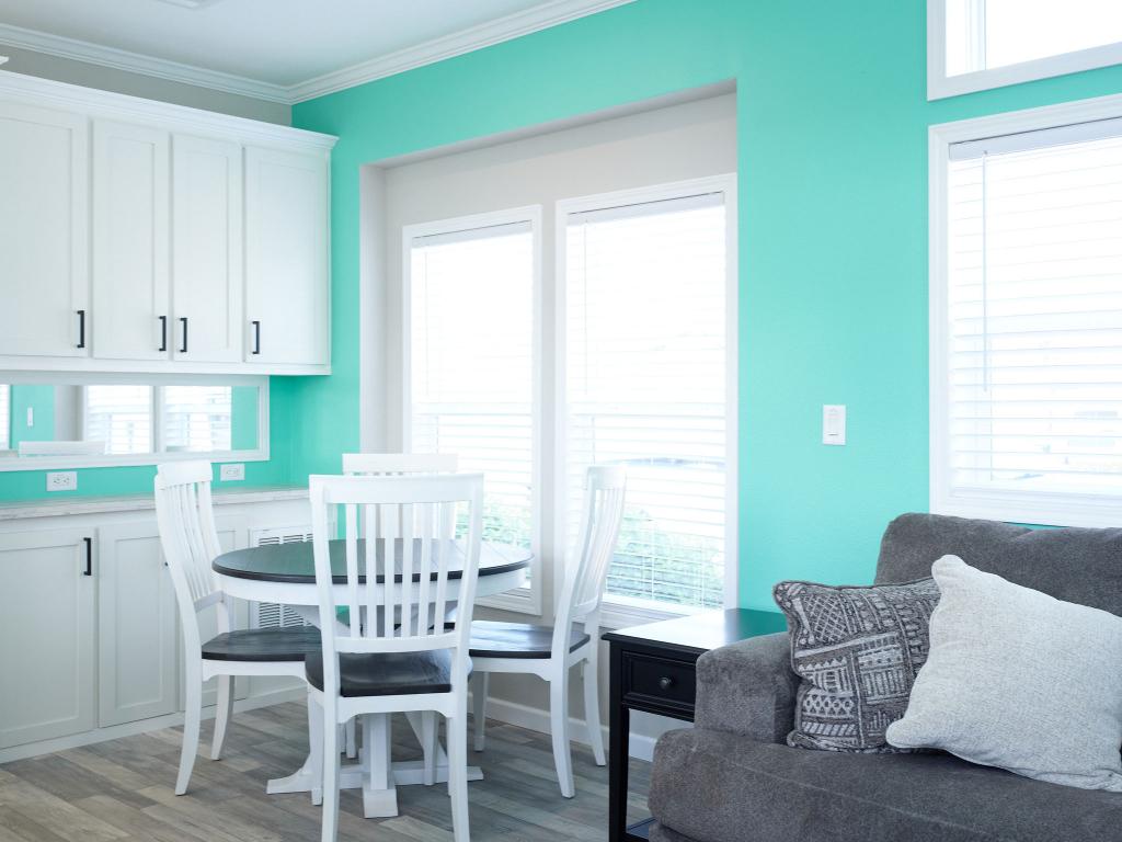 Bright kitchen with turquoise walls, white cabinets, a round dining table with four chairs, and a gray sofa with patterned cushions. Large windows let in natural light.