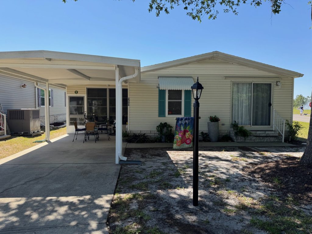 Single-story manufactured home with a carport, patio furniture, a small garden flag, and a lamp post in the front yard. The house has white siding and large front windows.