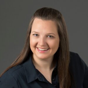 A woman with long straight brown hair, wearing a dark button-up shirt, smiles at the camera against a plain gray background.