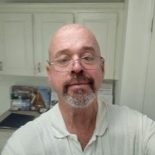 A middle-aged man with glasses, a bald head, and a white beard takes a selfie in a kitchen with white cabinets in the background.