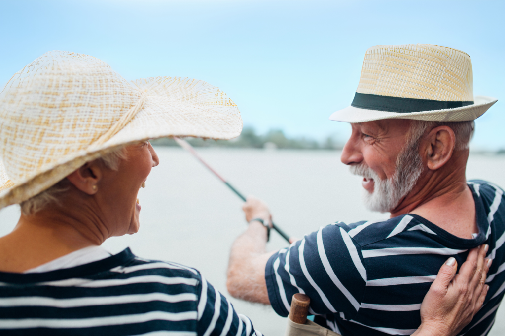 Two older adults wearing striped shirts and straw hats smile and fish together outdoors, with a blurred water background.