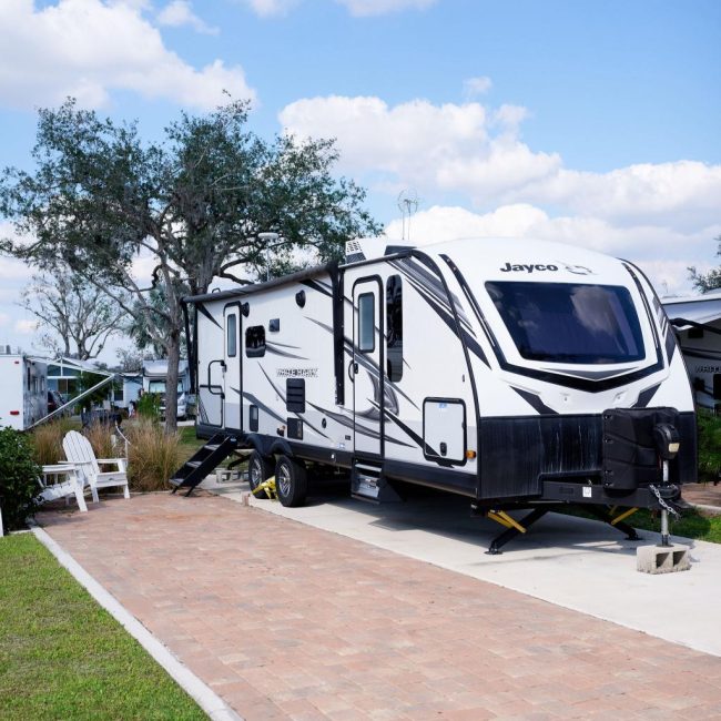 White Jayco travel trailer parked on paved area with steps extended, surrounded by grass and trees under a blue sky with clouds. Two white chairs sit nearby.