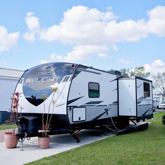 A white and black Twilight Signature trailer is parked on a concrete pad. There are two potted plants in front of it and grass surrounding the area. The sky is blue with scattered clouds.