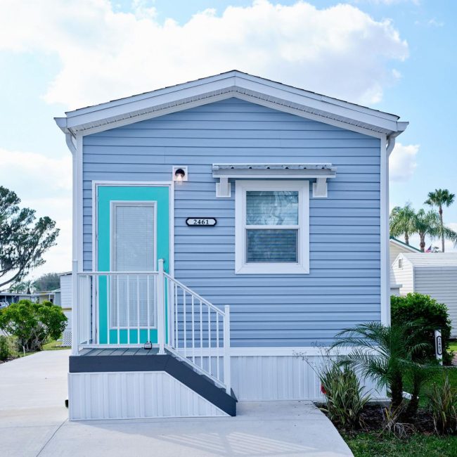 A small blue mobile home with white trim and a teal door. A short stairway leads to the entrance. There are a few plants in the foreground and trees in the background against a cloudy sky.